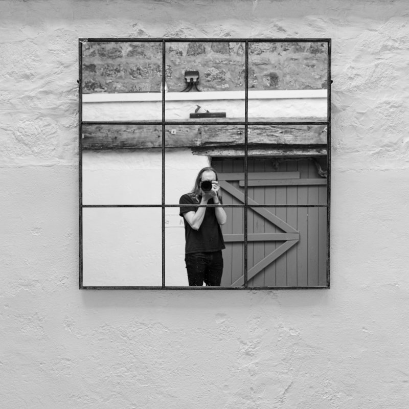 A photo of a mirror divided into a 3 by 3 grid, like a tic-tac-toe (or naughts and crosses) grid. The mirror is hung on a white wall, and in the mirror you can see an out of focus photographer. Behind the photographer is a wall, and mounted on that wall is an old block style wood plane.