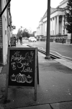 A chalk sign on a pavement board saying "Pumpkin Spice Latte" with illustration of pumpkins and a coffee cup. The street behind is in soft focus, but you can make out people walking and a car, and in the background the columns of an old grand building.