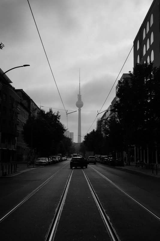 A photo of a look down a Berlin street with the tall spire of the Fernsehturm TV tower at the end of it. The tower is a giant concrete needle like structure with a ball on it about two thirds of the way up. The road is lined with tall buildings on both sides, and has two pairs of tram lines leading along it towards the distant tower, and an SUV is just pulling into the street ahead of us.