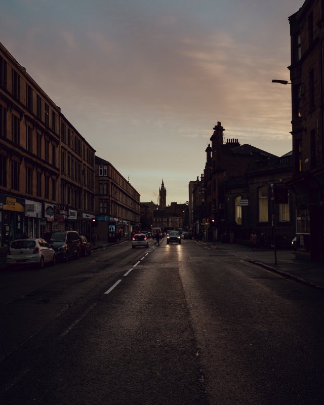 The view along a road lined with red-sandstone tenement buildings in the early morning, with the sky faintly blue with a little peach from the sunrise. At the end of the road you can see the profile of a tall old spire that is the main building of the University of Glasgow.