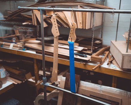 A photo of two guitar necks hung up on rack in room otherwise filled with wood on shelves. Both necks are made from maple: one is hung so you can see the back, showing the birds-eye figuring, and the other is hung so you can see the fretbaord is covered in masking tape.