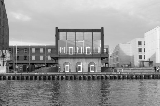 A square shaped building with mostly glass frontage sits on a harbour front, as seen from the river.