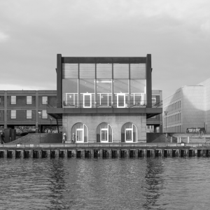 A square shaped building with mostly glass frontage sits on a harbour front, as seen from the river.