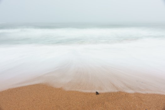 A long-exposure photo take on a beach looking straight out to sea, with the blurred waves mixing into the fog filled sky.