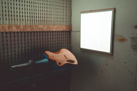 A moodily lit photo of a guitar body on a stand in a spray-booth, lit by a white square light panel. The guitar body was orange and is now sanded to show the wood through the colour.