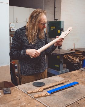 A photo of me stood behind a workbench in a workshop, I'm holding a guitar neck in my hands examining it, and on the workbench in front of me are two other guitar necks. Behind me is a bandsaw and a bin full of offcut bits of wood.