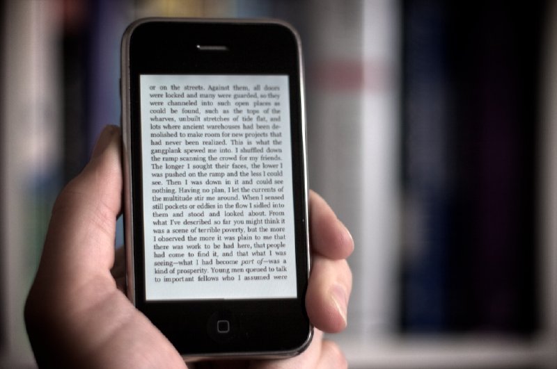 A photo of a hand holding an iPhone showing a page from an ebook. In the background, out of focus, is a wall of filled bookshelves.