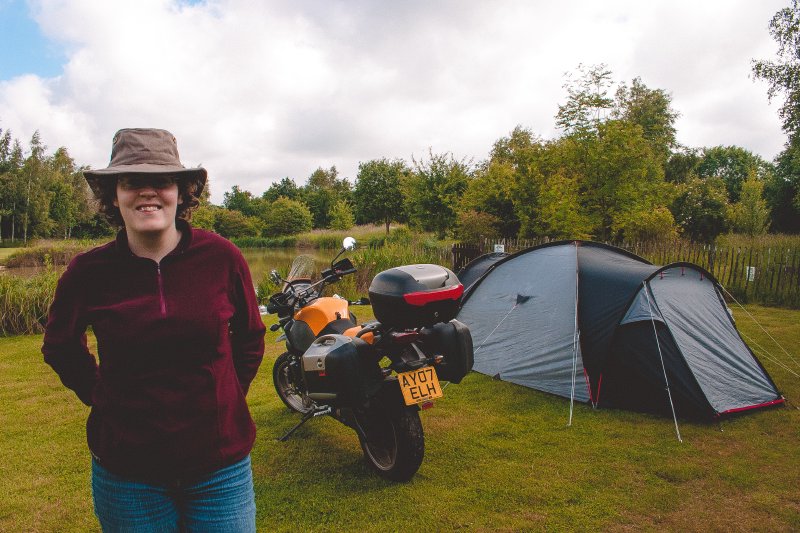 A smiling Laura stood in a green camp site next to a tent and a motorbike.