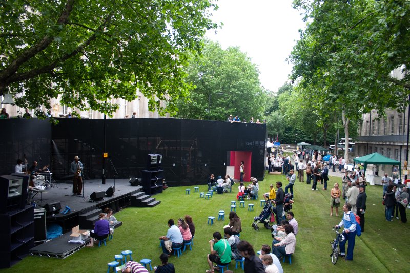A stage with musicians playing to people watching sat on real grass laid over road surface in central London.