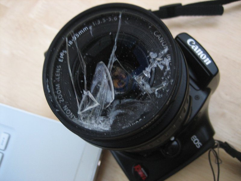A photo of a DSLR sat on a desk, and the front of the lens on the camera is all smashed glass.