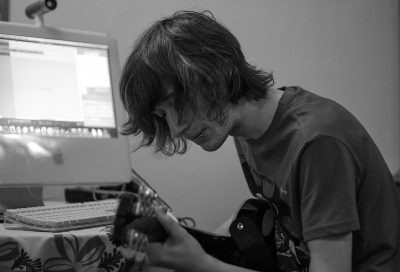 My brother sat playing guitar, whilst in the background an iMac looks on.