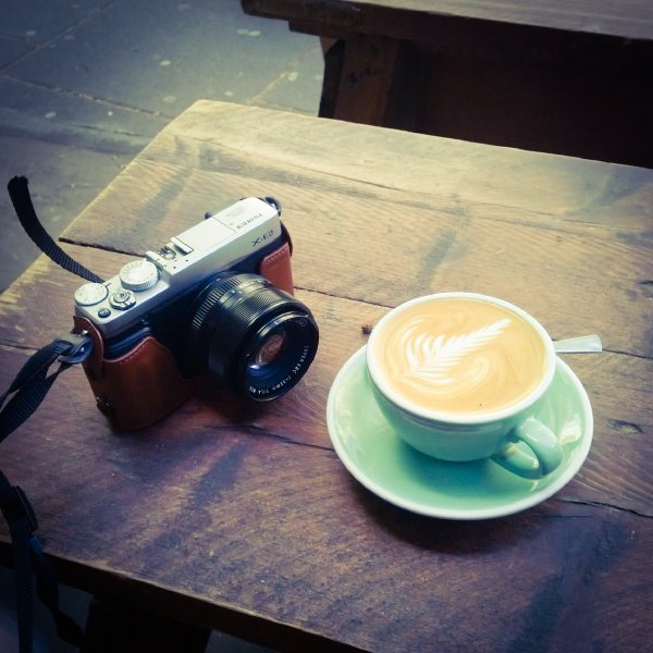 A shot of a small retro-styled digitial camera, the Fujifilm X-E2, sat on a table next to a Flat White at FreeState.