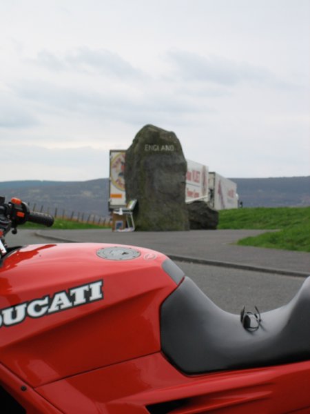 The fuel tank of the red Ducati in the foreground with a large stone with England written on it in the background.