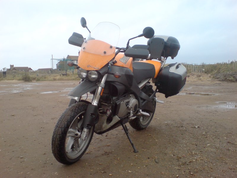 A muddy looking orange motorbike parked in a sandy carpark.