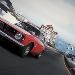 A screenshot from Forza showing a red classic Alfa Romeo racing along the start/finish straight of a track set in the mountains.