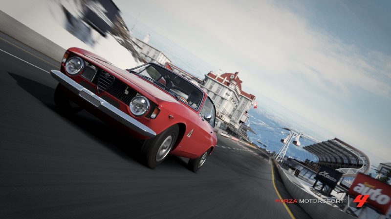 A screenshot from Forza showing a red classic Alfa Romeo racing along the start/finish straight of a track set in the mountains.