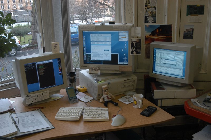 A picture of a desk with 3 CRT monitors on it (well, 2 on the desk, and a third on stack of binders on a chair). 