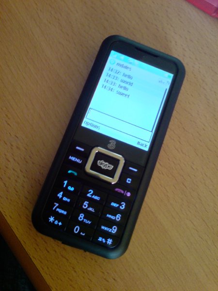 A picture of a candy-bar style mobile phone on a desk, which has the 3 network logo on it and a skype logo on one button.