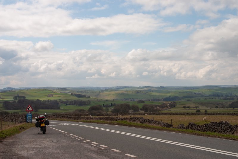 A photo of a green rolling countryside taken from a road lay-by, in which you can see parked a motorbike with panniers.