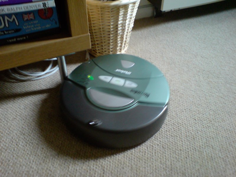 A blurry shot of a green disk shaped robot on the floor in a living room.