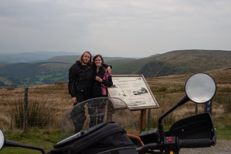 A picture taken from the camera balanced on the motorbike so you can see the screen and handlebars, looking at me and Laura stood by a viewpoint sign and the view of rolling hills.