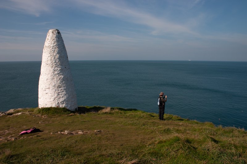 Laura stood by a cliff, looking out over the sea. Next to here is a large white stone cairn.
