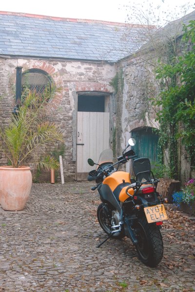 A photo of an old brick courtyard with a bright big yellow motorbike parted in it.