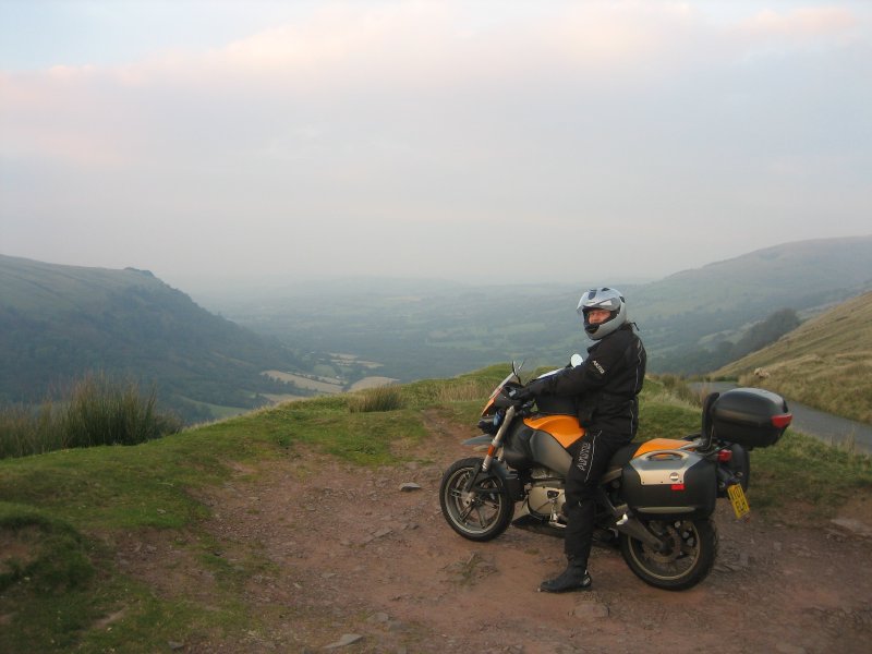 A picture down a green valley from atop one side, with me sat on my motorbike looking at the photographer.