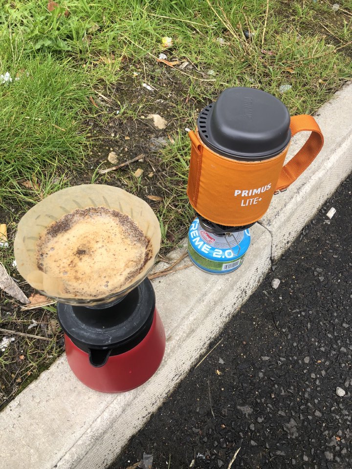 A small single cup primus gas burner and a V60 coffee brewer sit on a road curb next to some grass.