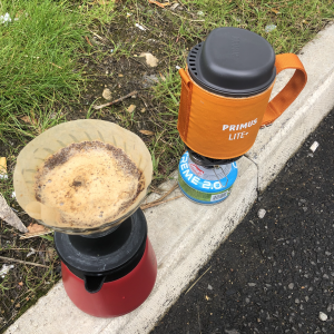 A small single cup primus gas burner and a V60 coffee brewer sit on a road curb next to some grass.