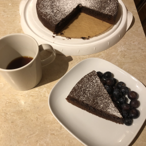 A photo of a slice of kladdkaka, Swedish chocolate mud cake, on a plate with some blueberries. Next to it is a cup of black coffeee, and behind them is the entire kladdkaka, minus two slices.