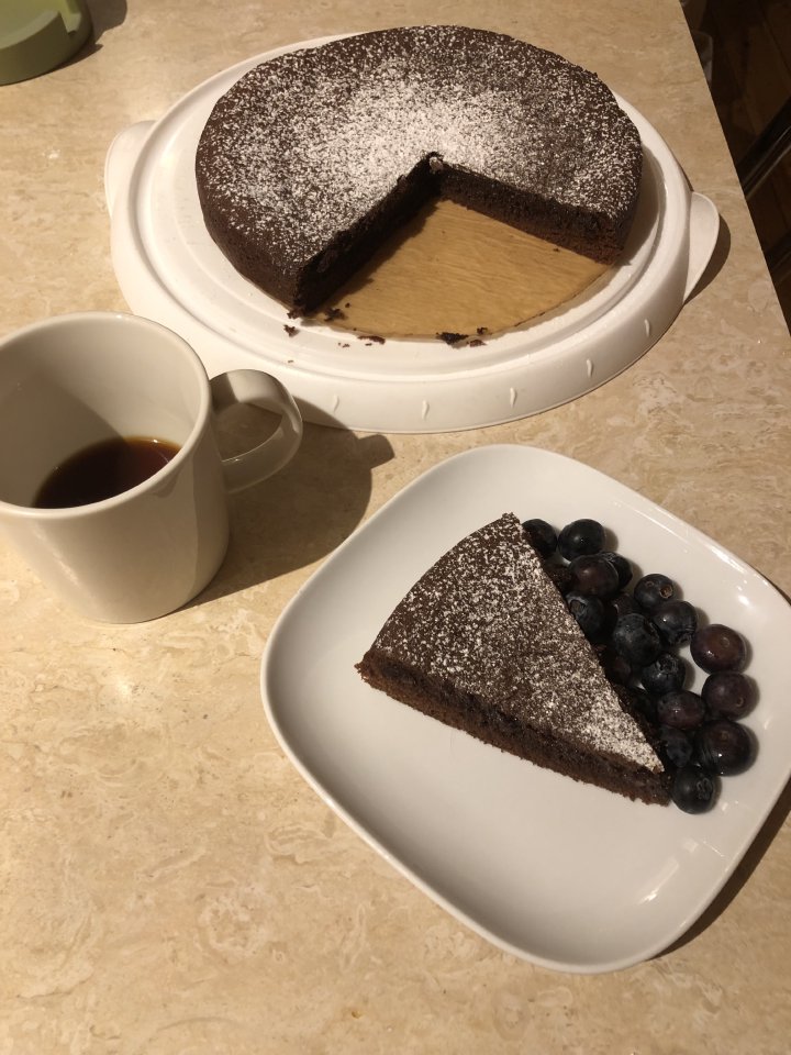 A photo of a slice of kladdkaka, Swedish chocolate mud cake, on a plate with some blueberries. Next to it is a cup of black coffeee, and behind them is the entire kladdkaka, minus two slices.