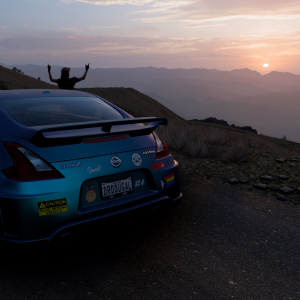 A in-game photo from Forza Horizon 5, showing a Nissan 370Z covered in stickers parked up on a mountain top at sunrise, with a driver stood behind the car watching the sun.