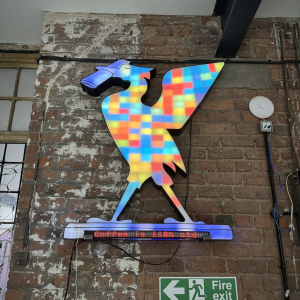 A photo of a large wall mounted display in the shape of the Liverpool liver bird, lit up in all manner of colours. Under the bird is an LED banner that reads 'Coffee is 119h old'