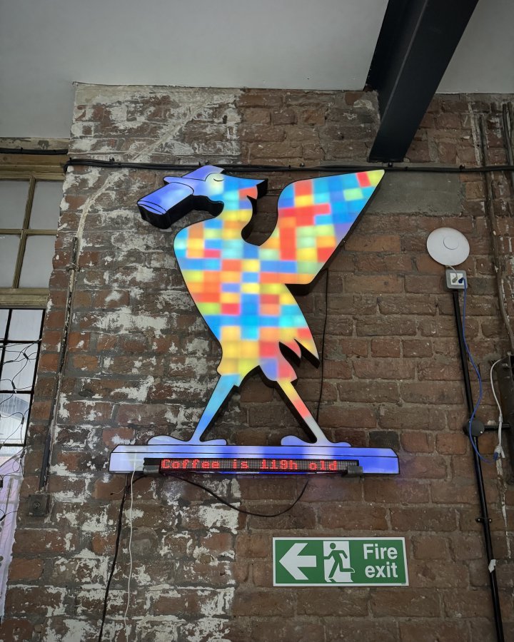 A photo of a large wall mounted display in the shape of the Liverpool liver bird, lit up in all manner of colours. Under the bird is an LED banner that reads 'Coffee is 119h old'
