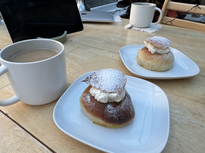 En foto av två semlor på små vita tallrikar, bredvid två kopp te.