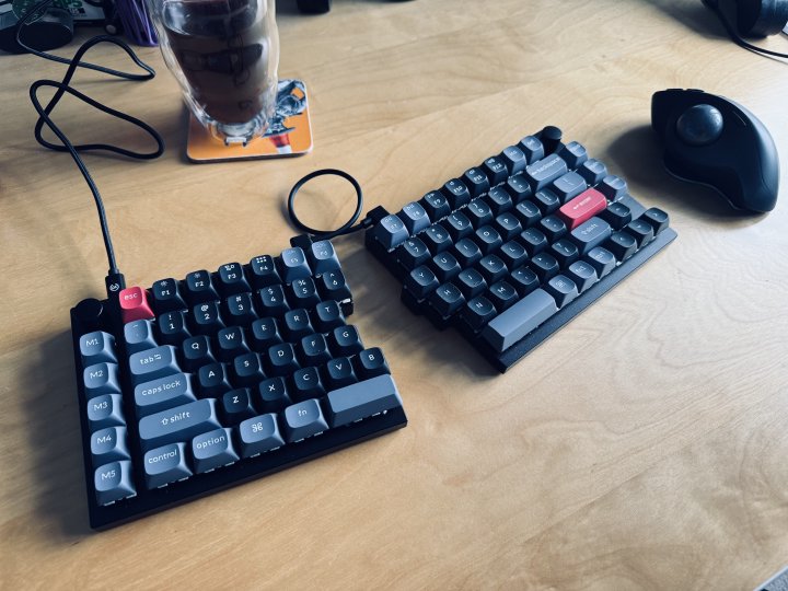 A photo of a split mechanical keyboard, in black with a mix of black, blue, and red keycaps. It sits on a desk, next to the keyboard is a trackball and a cup of coffee.