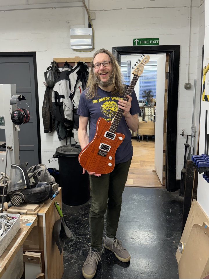 A photo of me in a workshop holding an orange electric guitar, smiling. The guitar is made from wood stained a bright orange with dark grain patterns, and has a maple neck with a rosewood fretboard.