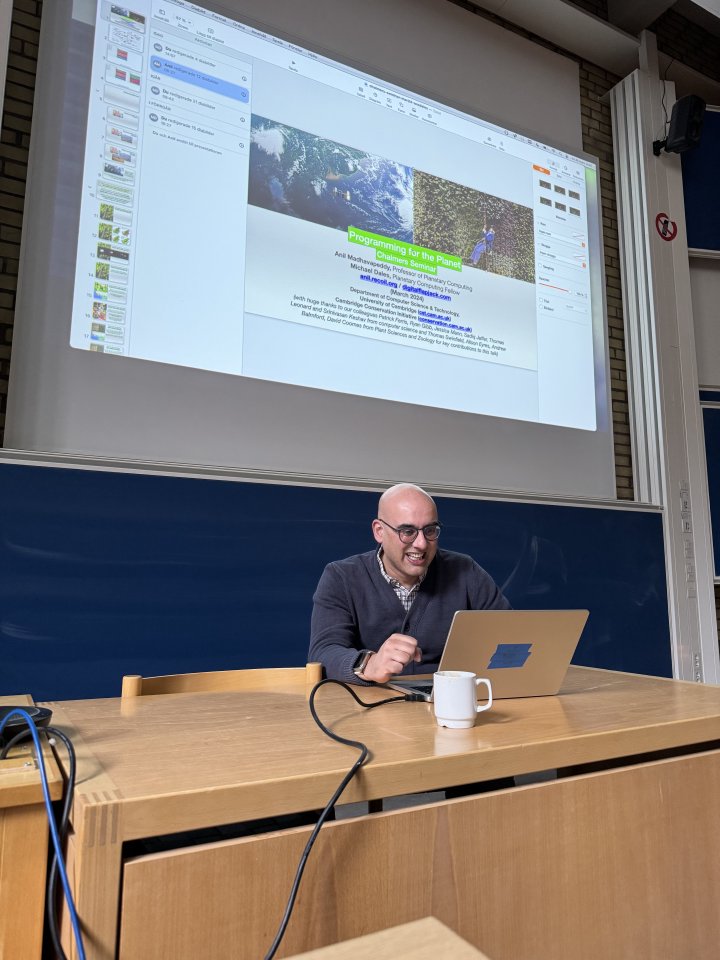 A photo of Anil sat at a desk at the front of a lecture theatre with a laptop, and behind him is a projection of a title slide of a talk `Programming for the planet` with both his name and mine as presenters.