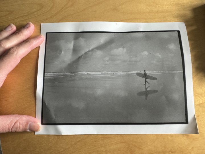 A photo of me holding a photo flat on my desk. The inner photo is a grainy black and white shot of a surfer walking to the water with their board.
