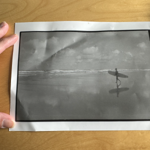 A photo of me holding a photo flat on my desk. The inner photo is a grainy black and white shot of a surfer walking to the water with their board.