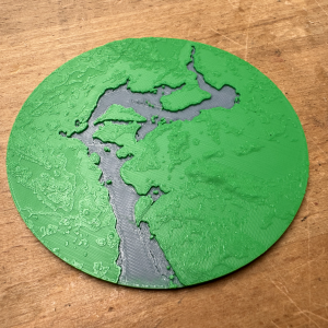A photo of a circular 3D-printed map section, showing a river with some islands in it. The river is printed in blue and recessed into the landscape, and the ground is printed green with some texture to indicate height differences.