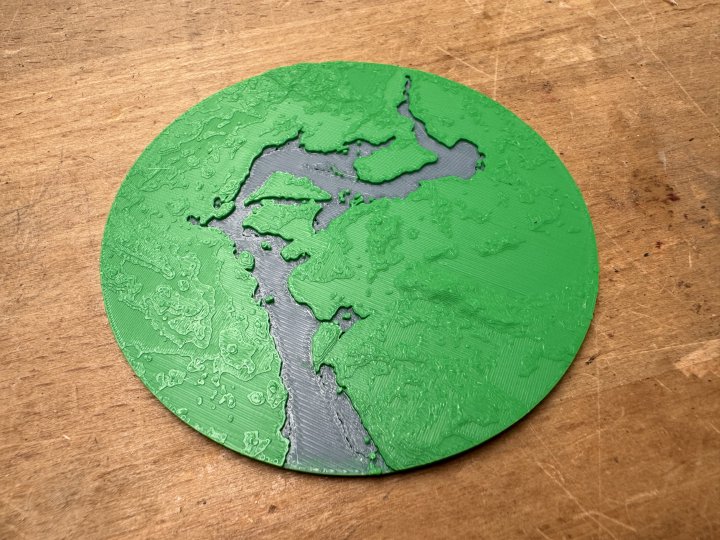 A photo of a circular 3D-printed map section, showing a river with some islands in it. The river is printed in blue and recessed into the landscape, and the ground is printed green with some texture to indicate height differences.