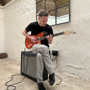 A photo of my brother sat on a large twin speaker guitar amplifier playing an orange electric electric guitar that has my signature on the headstock.