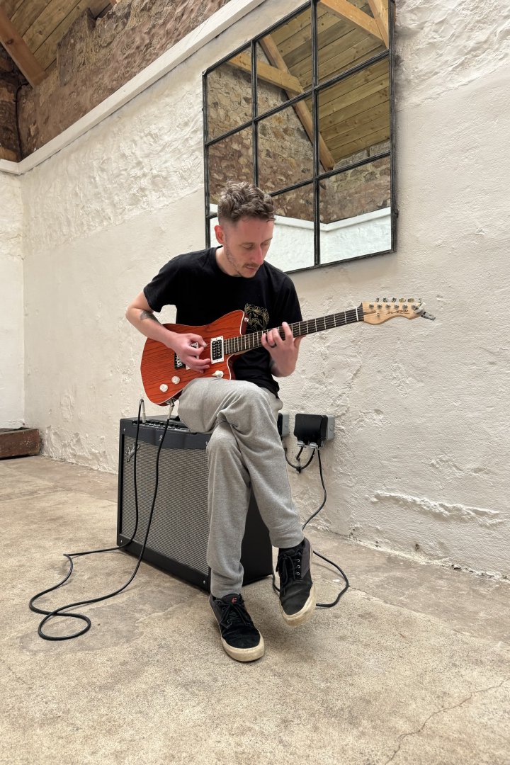 A photo of my brother sat on a large twin speaker guitar amplifier playing an orange electric electric guitar that has my signature on the headstock.