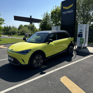 A photo of a yellow smart EV parked at a Fastned charging station in the sun. In the background, about 50 metres away and surrounded by trees, you can make out The Angel of the North, the large sculpure by Anthony Gormley just outside newcastle.