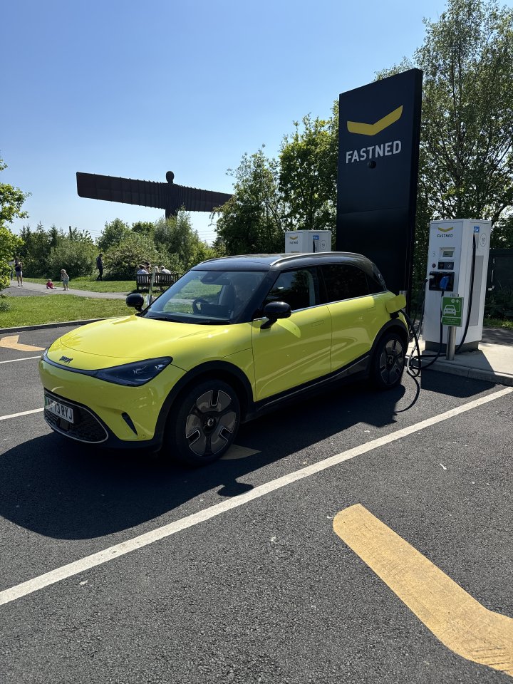 A photo of a yellow smart EV parked at a Fastned charging station in the sun. In the background, about 50 metres away and surrounded by trees, you can make out The Angel of the North, the large sculpure by Anthony Gormley just outside newcastle.