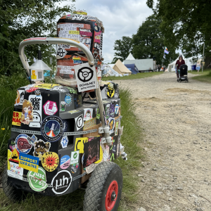 A photo of a small trolly consisting of some speakers stuck together with a canister and totally covered in silly stickers. It's sat at the side of a trail as someone pushes a baby buggie the other way.