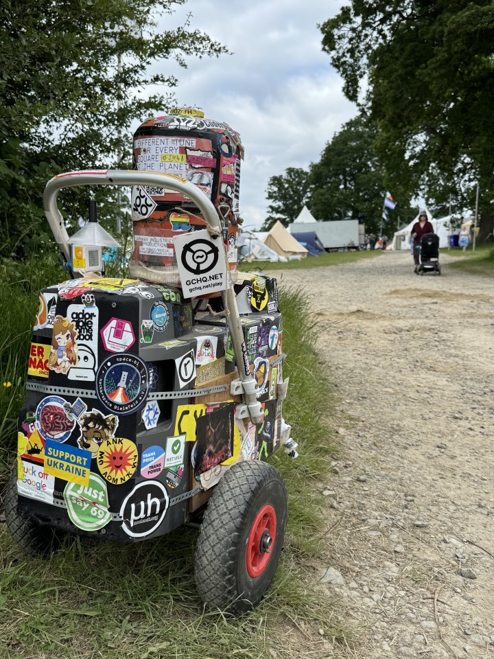 A photo of a small trolly consisting of some speakers stuck together with a canister and totally covered in silly stickers. It's sat at the side of a trail as someone pushes a baby buggie the other way.