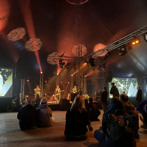 A photo of a stage in a large tent, on which three band members are performing, pressed in mokish gold robes. The left most is playing a laptop (doing the visualisations that are projected either side of the stage), in the middle is a woman singing whilst playing a Gibson SG guitar, and on the right a man is playing bass guitar. In front of the stage is a small chill crowd enjoying the show.